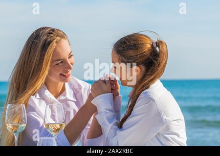 Zwei schöne junge Frauen halten die Hände und schauen sich mit Zuneigung auf einen außer Fokus gernden Meereshintergrund an. Romantisches Konzept. Stockfoto