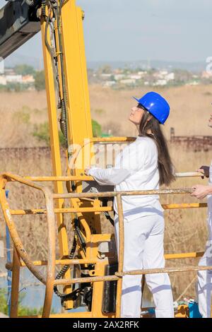 Schön fokussierte Frau mit Schutzbrille und Helm, die das Bedienfeld eines Teleskopauslegers auf einem Hintergrund mit unsichtlichem Fokus bedienen. Zusammenarbeit Stockfoto