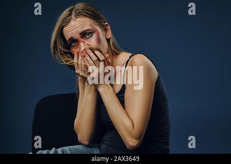 Blonde Frau in schwarzem Unterhemd und Jeans. Blutendes Gesicht mit Prellungen, sie bedeckt es mit Haaren und Handflächen, auf blauem Hintergrund sitzend. Tun Stockfoto