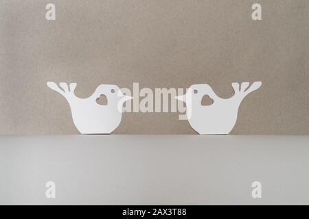 Zwei weiße, papiergeschnittene Vögel mit gegeneinander sitzenden Herzen auf einem weißen Pappsockel auf beigefarbenem Hintergrund. Valentinstag und Liebeskonzept Stockfoto