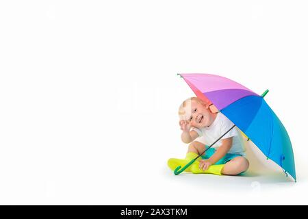 Ein blondes Kind kaukasisch sitzt unter einem regenbogenhellen Regenschirm in Gummistiefeln und Wellen seine Hand auf einem weißen Hintergrund. Konzept über Puddel, Sicherheit Stockfoto