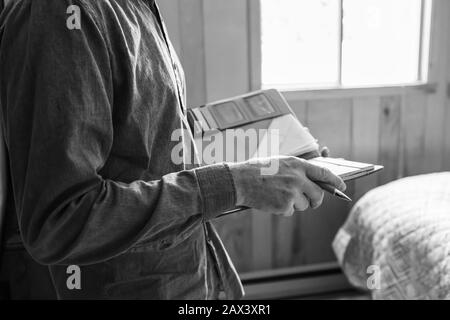 Seitliche Nahaufnahme und selektive Fokussierung Schwarz-Weiß-Sicht auf die Hände des Mannes, während er Stift und Notizbuch mit Lederkarten-Halter-Abdeckung, Schlafzimmer-Hintergrund hält Stockfoto