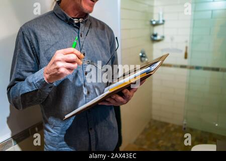 Der Inspektor spricht und hält ein Notizbuch, einen Stift und seine Brille, ein kaukasischer Mann, der mit Badezimmer und Duschkabine im Hintergrund aufschließt. Stockfoto