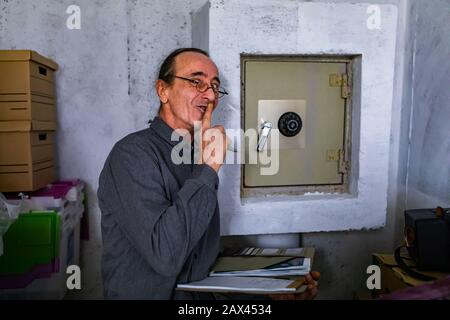 Hausinspektor während einer Inspektion, Mann mittleren Alters, der neben dem geheimen Metall-Safe an der Wand steht, blickt er mit dem Finger auf Lippengeste zur Kamera Stockfoto