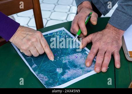 Alte Erwachsene Hände auf Holztisch nah oben, die Hand der Frau zeigt auf die Lokalisierung auf einer Papierkarte neben der Hand des Mannes mit einem Stift, während er ein Auge darauf behält Stockfoto