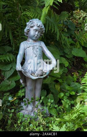 Nahaufnahme der blauen Polyresin Statue von jungen Mädchen in der Grenze Mit Petasiten - Butterbur Pflanzen im privaten Garten im Garten Sommer Stockfoto