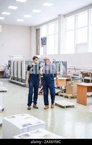 Leitende und junge Mitarbeiter der Druckhäuser in blauen Overalls, die die moderne Werkstatt durchqueren und über die Arbeit diskutieren Stockfoto