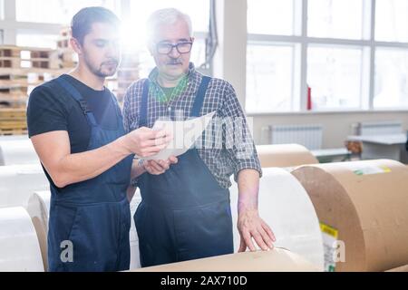 Ernsthafte Mitarbeiter von Druckhäusern in Overalls, die Notizen in Papier anzeigen, während sie Papierrollen im Lager zählen Stockfoto
