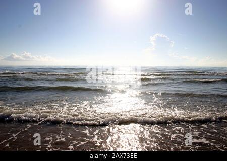 Sonnenlicht funkelnd, wie es auf die sanften Wellen reflektiert, die an einem sonnigen Nachmittag auf einen sandigen Strand rollen. Stockfoto
