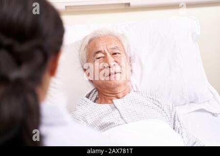 Asiatischer alter Mann, der im Bett liegt und mit junger Ärztin in der Krankenstation spricht Stockfoto