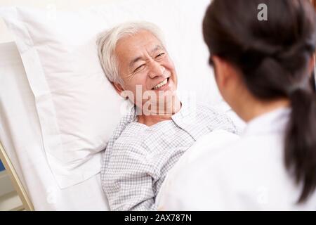 Asiatischer alter Mann, der im Bett liegt und mit junger Ärztin in der Krankenstation spricht Stockfoto