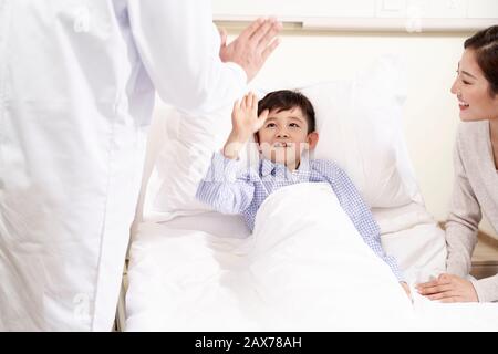 Glückliches fünf Jahre altes asiatisches Kind, das im Bett in der Krankenstation liegt und dem Arzt einen Hi-Five gibt Stockfoto