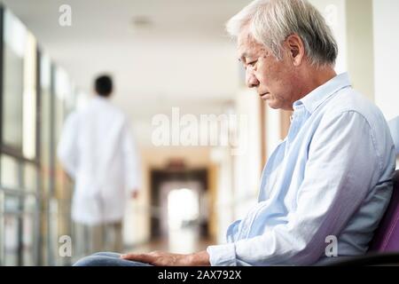 Asiatischer alter Mann, der auf dem Flur des Krankenhauses sitzt, sieht traurig und depressiv aus Stockfoto