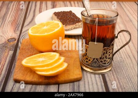 Tee aus facettiertem Glas in einem Vintage-Cup-Halter, geschnittene Zitrone auf einem Brett und ein Stück Waffelkuchen auf einem weißen Teller auf Holzhintergrund. Nahaufnahme Stockfoto