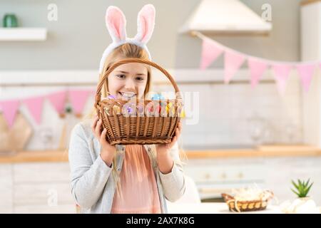 Porträt des lustigen kleinen Mädchens, das am Tisch sitzt und mit Hünenohren trägt und den Osterkorb mit Eiern hält Stockfoto
