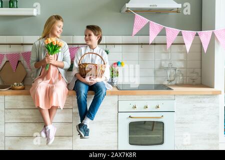 Portrait von kleinen Jungen und Mädchen, die in der Küche sitzen. Sie halten einen Korb mit Ostereiern und Blumenstrauß mit Tulpen Stockfoto