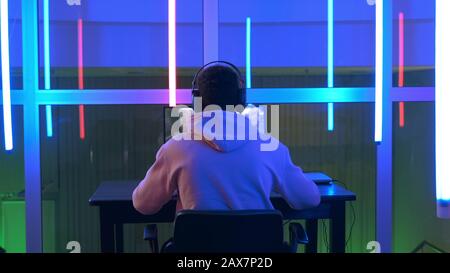 Professioneller Mixed-Race-Spieler, der in Shooter Online-Videospiel auf dem pc in einem großen Raum mit warmfarbigen LED-Lichtern spielt. Stockfoto