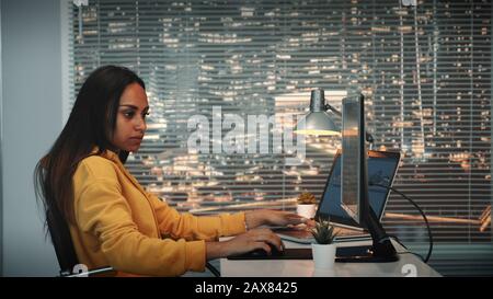 Seitenansicht des schwarzen weiblichen Videofilmers, der mit Filmmaterial und Ton auf dem Computer mit zwei Displays arbeitet und Videos aus verschiedenen Bildern einstellt Stockfoto