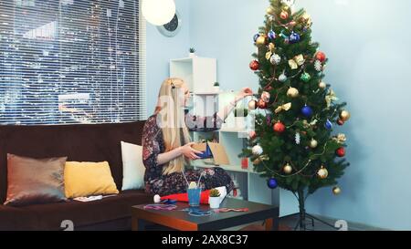 Hübsches Mädchen hängt Spielzeug am Weihnachtsbaum mit Wolkenkratzern auf dem Hintergrund und sitzt auf einem Sofa mit Kopfkissen zu Hause. Sie nimmt die Spielzeuge aus der Schachtel. Stockfoto