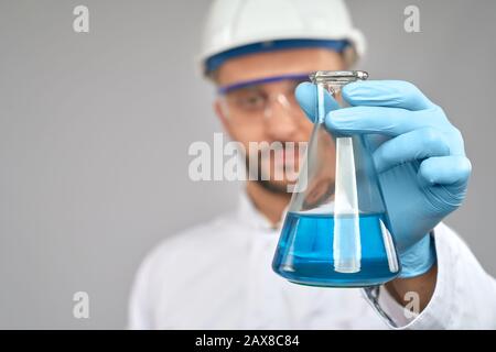 Nahaufnahme des jungen Chemikers in Brille und Helm, der im Labor experimentiert, isoliert auf Grau. Selektiver Fokus von Kolben mit blauer Flüssigkeit in der Hand des Wissenschaftlers in Handschuhen und weißem Labormantel. Stockfoto