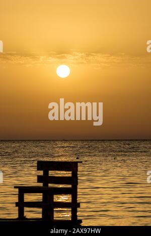 Konzentrieren Sie sich auf die runde Einstellung Sonne. Verschwommene Sitzbank Silhouette am Strand bei Sonnenuntergang vor goldenem Ionischen Meer Wasser in der Nähe der Küste. Abenddämmerung aus Ksamil, Albanien, Frühlingslandschaften. Stockfoto