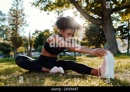 Lächelndes Porträt einer sportiv sitzenden afroamerikanischen jungen Frau auf dem Rasen, die ihre Beine im Park streckt - fröhliche junge schwarze Frau, die sich aufwärmt Stockfoto