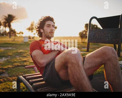 Fit Young man hört Musik auf Ohrhörern, die ABS-Übungen auf der Bank im Fitnesspark machen - motivierter Mann, der in einem Outdoor-Fitnessstudio abknirscht Stockfoto