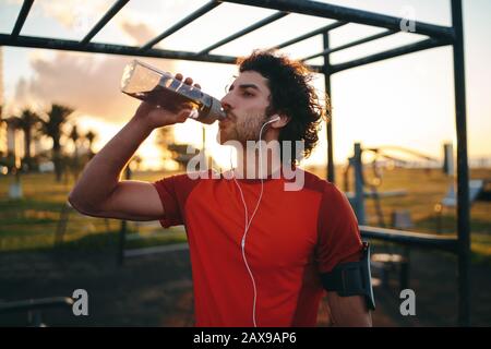 Sportlicher junger kaukasischer Mann, der Musik auf Ohrhörern hört, die Wasser aus einer transparenten, wiederverwendbaren Flasche im Sportpark im Freien trinken Stockfoto
