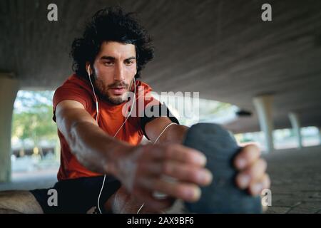 Portrait eines gesunden Sportlers, der sich vor dem laufen unter der städtischen Brücke ausdehnt und aufwärmt - Nahaufnahme eines ernsthaften männlichen Läufers Stockfoto