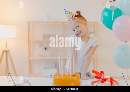Kleines Ingwermädchen trägt Partyhut und weißes Tshirt, steht in der Nähe eines Festtisches mit Kuchen, bläst Kerzen und macht sich während ihres Geburtstags Wunsch, posiert in w Stockfoto