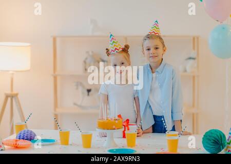 Froher Junge und seine kleine Ingwerschwester, die in festliche Kleidung gekleidet sind, feiern gemeinsam Geburtstag, umgeben von Kuchen, Geschenk und Tassen von dr. Stockfoto