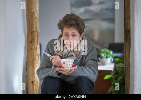 Junger kranker Mann, der in seinem Haus mit einer Tasse Tee und einem Thermometer in den Händen sitzt Stockfoto