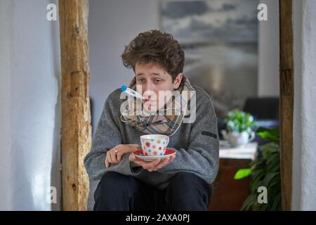 Ein kranker junger Mann mit einer Kälte, der in seinem Haus sitzt, hat ein Thermometer im Mund und hält einen Becher in der Hand Stockfoto