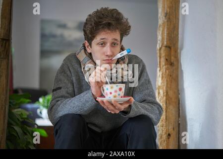 Ein kranker junger Mann mit einer Kälte, der in seinem Haus sitzt, hat ein Thermometer im Mund und hält einen Becher in der Hand Stockfoto