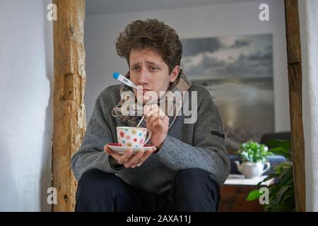 Ein kranker junger Mann mit einer Kälte, der in seinem Haus sitzt, hat ein Thermometer im Mund und rührt eine Tasse Tee mit einem Löffel Stockfoto