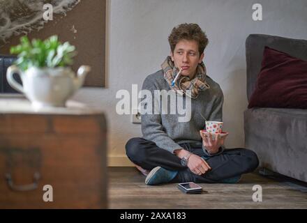 Junger depressiv aussehender Mann mit einem kalten Sitzen auf dem Boden in seiner Wohnung mit einem Thermometer im Mund und einer Tasse Tee in der Hand Stockfoto