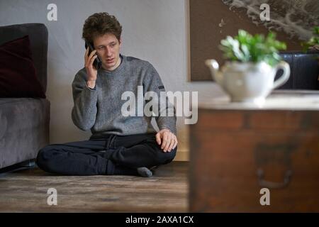 Junger Mann sitzt auf dem Boden in seiner sonnigen Wohnung und spricht über ein Handy Stockfoto
