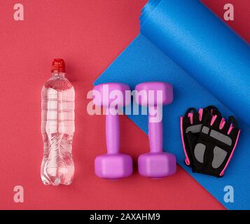 Wasserflasche, blau verdrehte Neopren-Matte, zwei Kurzhanteln und Handschuhe auf rotem Texturhintergrund, Draufsicht, Sportset Stockfoto
