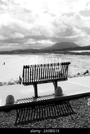 Eine Metallbank wirft einen Schatten mit einem abgeschiedenen Strand und Berg im Hintergrund. Magheraroarty, Co. Donegal, Irland Stockfoto