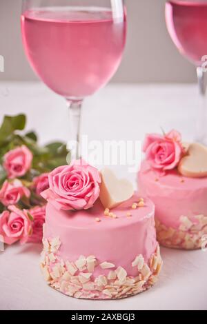 Zwei Minikuchen, eine Brille mit rosafarbenem Traubenwein und Rosenblumen auf einem weißen Tisch. Romantisches Dinner Konzept. Stockfoto