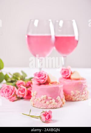 Zwei Minikuchen, eine Brille mit rosafarbenem Traubenwein und Rosenblumen auf einem weißen Tisch. Romantisches Dinner Konzept. Stockfoto