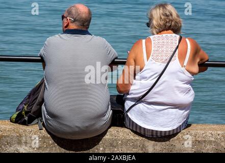 Ein übergewichtiges Paar im mittleren Alter, das an einer Meereswand sitzt und an einem Geländer am Meer festhält. Stockfoto