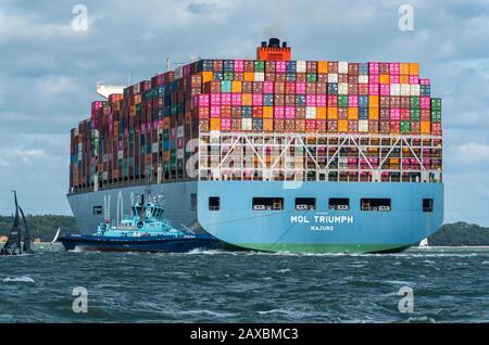 Ein großes Containerfrachtschiff, das in den Hafen von southampton eindringt, legt mit einem Schlepper an, der dazu beiträgt, das große massive Seeschiff in der solent zu steuern. Stockfoto