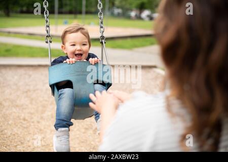 Mutter treibt fröhliches süßes Kleinkind auf dem Spielplatz in Schwung Stockfoto