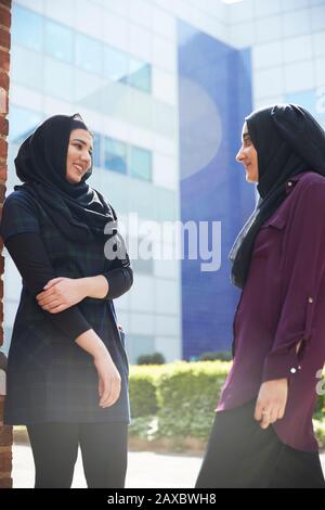 Junge Frauen Freunden sich in Hijabs an, die außerhalb des sonnigen Gebäudes sprechen Stockfoto