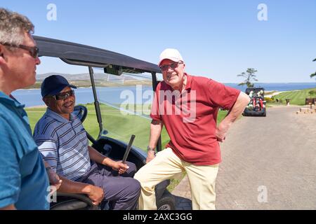 Glückliche männliche Golfspielerfreunde, die auf dem sonnigen Platz in der Golfkarre sprechen Stockfoto