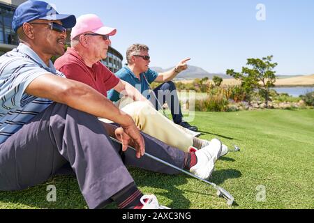 Männliche Golfer, die auf einem sonnigen Golfplatz im Gras sitzen Stockfoto