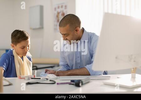 Ein männlicher Kinderarzt zeigt digitales Röntgenbild für Jungen mit Arm in Schlinge im Arztbüro Stockfoto