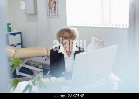 Portrait lächelnd, selbstbewusste Oberärztin, die am Computer in der Arztpraxis arbeitet Stockfoto