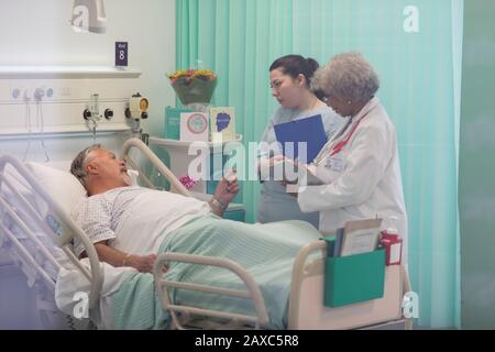 Ärzte mit medizinischen Diagrammen, die Runden drehen und mit dem leitenden Patienten im Krankenhauszimmer sprechen Stockfoto